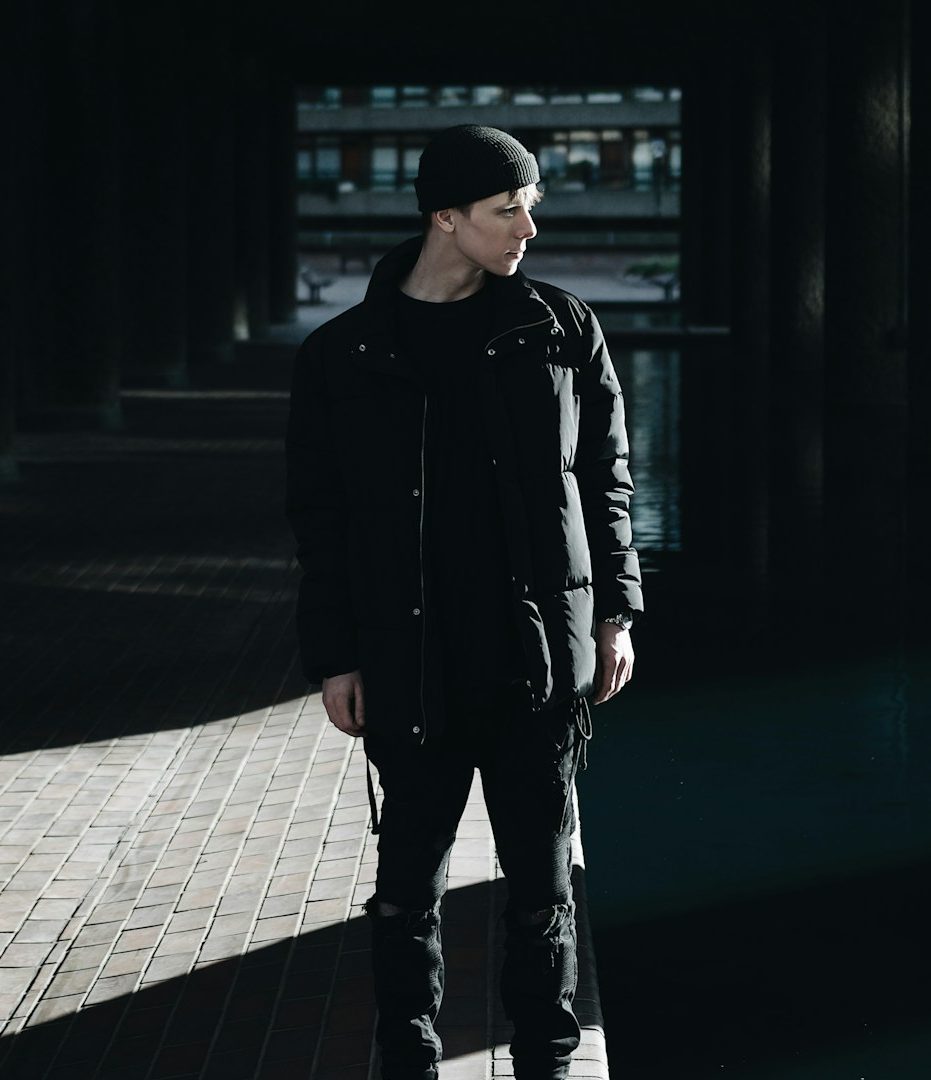 A person in a black outfit stands in a dimly lit urban setting with shadows.